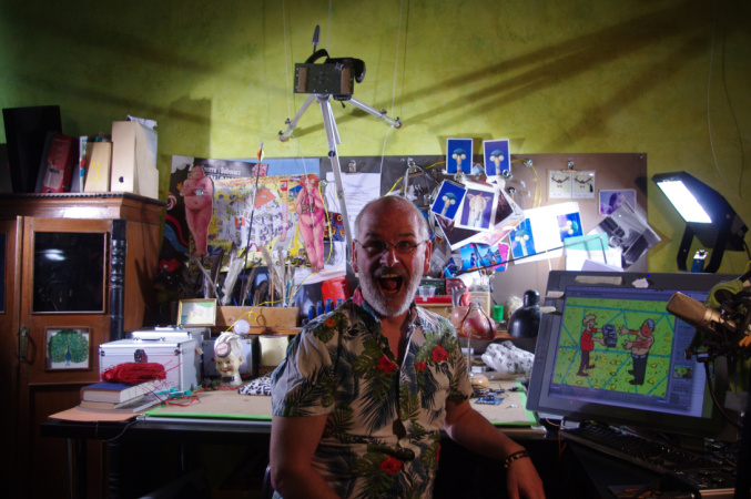 Friedel sitzt vor seinem Atelierschreibtisch. Im Hintergrund sind einige seiner Kunstwerke zu sehen.