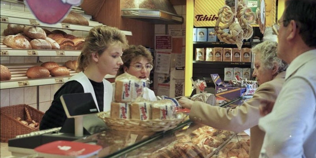 In einer Bäckerei stehen zwei Verkäuferinnen durch einen Tresen getrennt zwei Kunden gegenüber.
